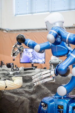 Der Interact Rover der ESA hat eine Gesteinsprobe eingesammelt und übergibt sie an DLR-Roboter Rollin‘ Justin. Auf dem Bildschirm im Hintergrund ist NASA-Astronaut Frank Rubio zu erkennen, der die Zusammenarbeit der Roboter von der Internationalen Raumstation ISS aus steuert. (Bild: DLR (CC BY-NC-ND 3.0))