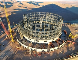 Dieses Bild, aufgenommen Ende Juni 2023, zeigt eine Drohnenaufnahme der Baustelle des Extremely Large Telescope der ESO auf dem Cerro Armazones in der chilenischen Atacama-Wüste. Dort sind Ingenieurinnen und Ingenieure, Handwerkerinnen und Handwerker gerade dabei, die Struktur der Teleskopkuppel in einem rasanten Tempo zu montieren. Die Stahlkonstruktion, die sich jeden Tag sichtbar verändert, wird bald die für Teleskopkuppeln typische runde Form annehmen. Die Menschen, die am unteren Bildrand zu sehen sind, geben dem Foto ein Gefühl von Größe und zeigen, wie groß die Kuppel des ELT sein wird. Hinter dem Teleskop sehen wir den Schatten des Cerro Armazones, der auf die Wüstenlandschaft projiziert wird. (Bild: ESO)