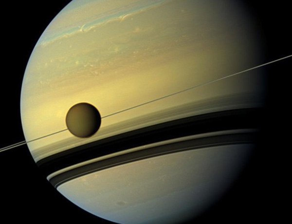 Saturn mit phänomenalen Wolkenbändern, die Ringe von links nach rechts des Bildes laufend sind als Strich fast unsichtbar, Titan als kleine braune Kugel davor